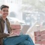 Young man sitting on a bench outdoors using a laptop, smiling at the camera.