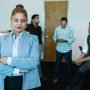 Confident businesswoman standing in modern office with colleagues in the background.